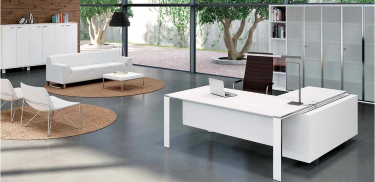Ambientazione ufficio con scrivania angolare dotata di piano in vetro temperato satinato retroverniciato bianco, box multiuso attrezzato con cassettiera portante, cassetto cancelleria con serratura, vano portaborse e top access con coperchio metallico. Sullo sfondo è presente una libreria alta modulare con 5 ripiani e ante in vetro satinato con telaio in alluminio brillantato.