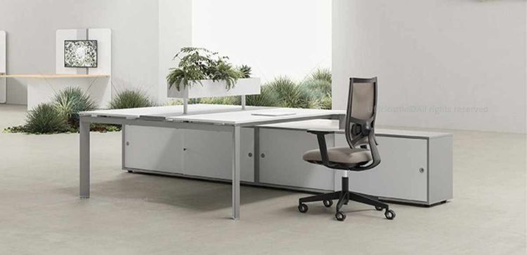 Bench a 2 postazioni composto da due scrivanie contrapposte con binario centrale attrezzabile in alluminio anodizzato, fioriera centrale, sportelli apribili per ispezione cablaggi, gambe metalliche a U rovesciata e due mobili di servizio portanti con ante scorrevoli, serratura a pulsante, piedini livellatori e finiture disponibili in melaminico colorato o neutro.